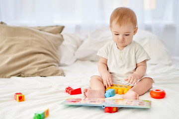 A little child sits on a bed with toys and a children's book.