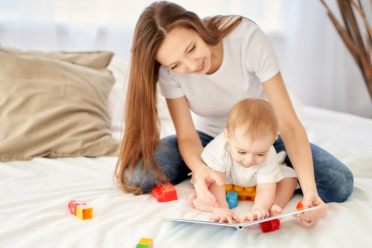 A young, beautiful mother with long hair sitting on the bed with her baby shows him pictures in a book.