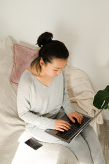 Asian woman dressed in grey shirt and trousers. Young Asian woman working from home while sitting on a sofa. Korean girl using laptop.