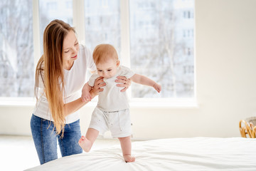 A young mother supports and teaches the baby to walk. The baby stomps on the bed.