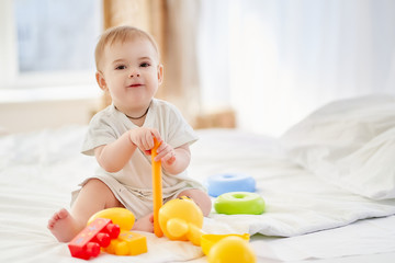 A little boy collects a toy pyramid sitting on the bed against the background of a blurry bright room.