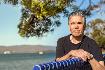 Portrait of a mature man 57 years with gray hair looking camera sitting on a bench at a beach outdoors, confident, relaxed.