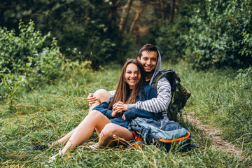 Naklejka na ściany i meble Woman with long hair and man sitting on the grass in the forest with backpacks. Have fun in nature, hugging and kissing