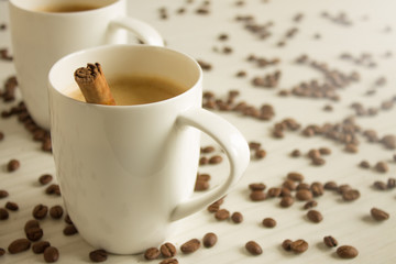 Two white coffee cups with a cinnamon stick on a wooden table with copy space and scattered grains