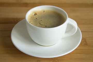 A small white Cup of fresh espresso with foam and a saucer close up on a brown wooden background