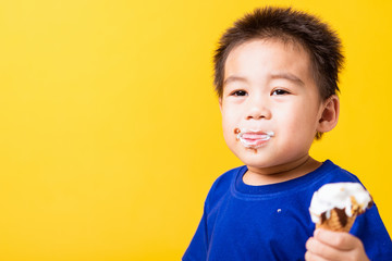 Happy portrait Asian child or kid cute little boy attractive laugh smile playing holds and eating sweet chocolate ice cream waffle cone, studio shot isolated on yellow background, summer concept