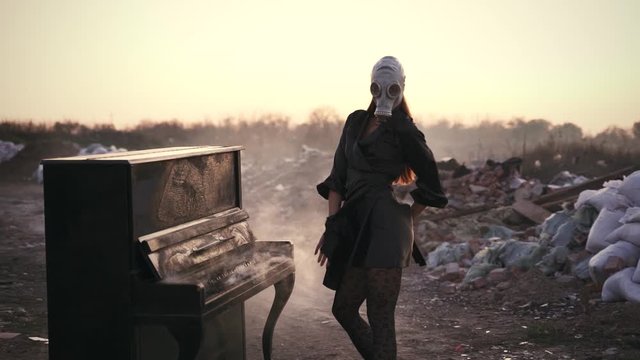 girl in a black dress, pantyhose and gas mask dancing near a smoking piano in a junkyard