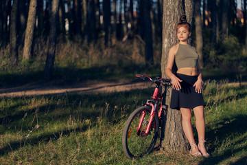 Fototapeta na wymiar A teenage girl in a top and skirt, standing with her eyes closed, leaned against a pine tree in the forest. Rest from cycling in nature.