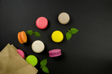 Multicolored macaroons and mint leaves on a black background, top view. Macaroons fall out of a paper bag