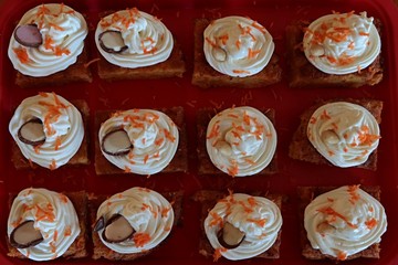 Carrot cakes decorated with whipped cream, sliced chocolate egg half with milk, strawberry flavoured fill or whole almond and grated fresh carrot on top. Top view, cakes are placed on plastic tray.