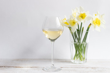 White wine in glass and yellow narcissus flowers in vase on white wooden background