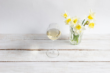 White wine in glass and yellow daffodils in vase on white wooden background