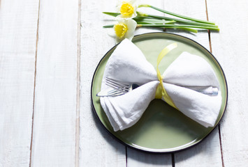 Knife and fork in white table napkin on green plate, daffodils on wooden background
