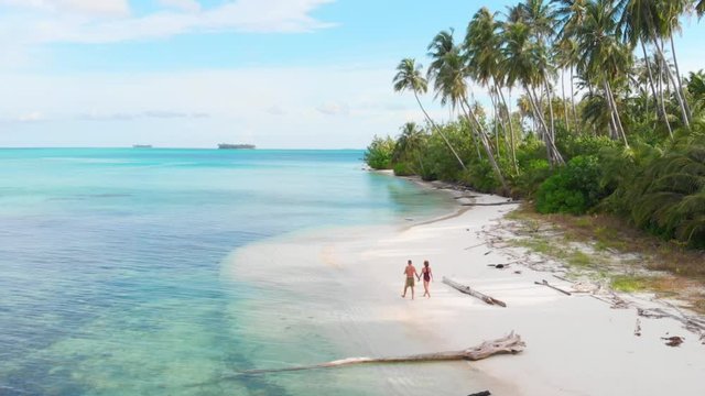 Aerial slow motion: couple walking on tropical beach at sunset, away from it all, caribbean sea white sand beach palm trees forest. Man and woman relaxing on perfect secluded island.