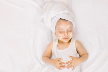 Fototapeta na wymiar Photo of relaxed pretty little baby girl with cream on her face and bath towel on head, lying with closed eyes on white background after spa bath procedures. Copy space
