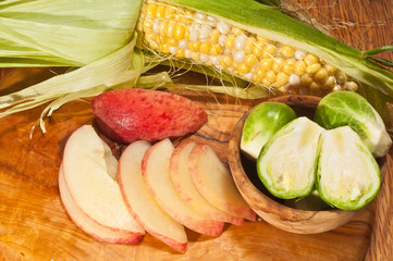 Top view, medium distance of a freshly picked, ear of corn striped to see kernels, ripe, local, peach, sliced in thin slices and rare wood bowl with Brussels sprouts, on a rare wood plate