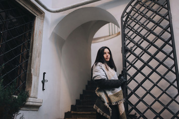 A young caucasian black-haired girl in a dark autumn coat and a wide scarf-wrap stands near black metal gate and looks straight. Lviv, Italian courtyard, Ukraine. Stay in place, quarantine concept.