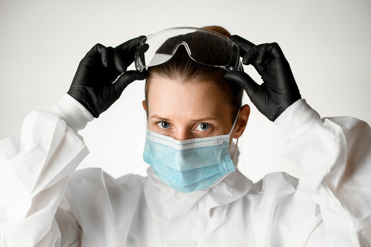 Young female nurse in medical mask and white protective suit adjusts goggles on her head