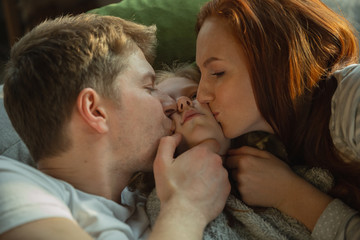 Kissing with all love. Family spending nice time together at home, looks happy and cheerful. Mom, dad and daughter having fun, lying down the sofa. Togetherness, home comfort, love, relations concept.