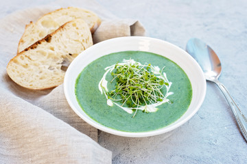 Cream soup of fresh spinach with cream and arugula microgreen in a white plate with ciabatta bread on a light gray background. Healthy and diet food. Flat lay.