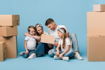Fototapeta na wymiar positive parents and kids holding frame and sitting on blue with cardboard boxes for relocation
