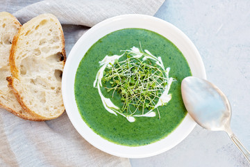 Cream soup of fresh spinach with cream and arugula microgreen in a white plate with ciabatta bread on a light gray background. Healthy and diet food. Flat lay.