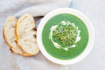Cream soup of fresh spinach with cream and arugula microgreen in a white plate with ciabatta bread on a light gray background. Healthy and diet food. Flat lay.
