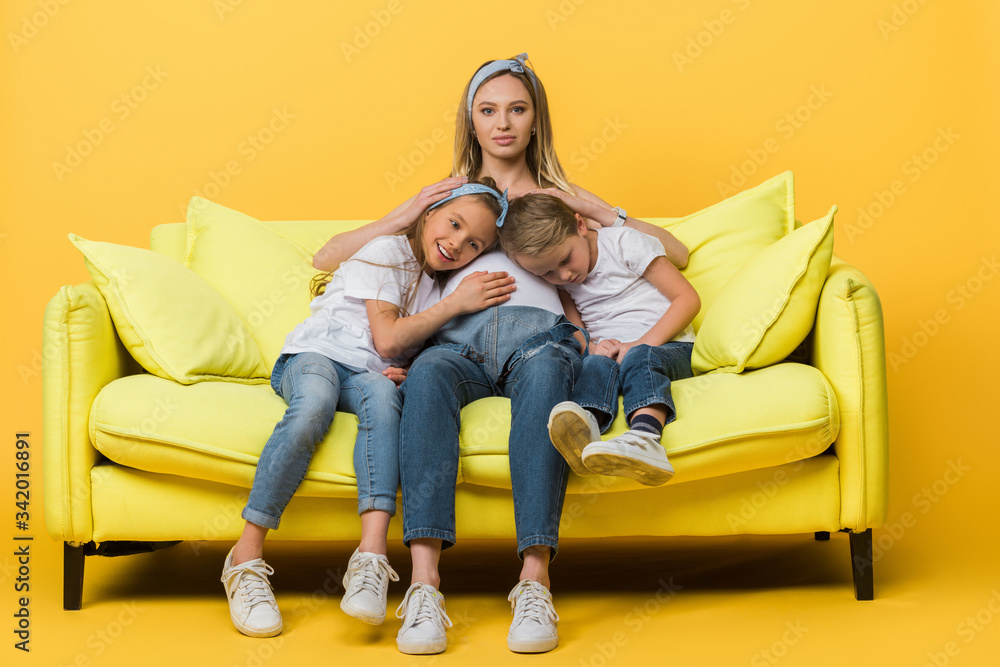 Wall mural pregnant mother hugging with smiling daughter and son on sofa on yellow - Wall murals