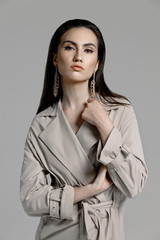 Beautiful sexy brunette woman in a black bodysuit, a beige trench coat with large gold earrings posing on a gray background in the studio. Close-up portrait of a model with professional liner make-up