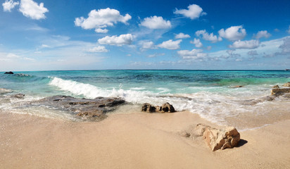 Fototapeta na wymiar Rocky beach view at Caribbean sea coast, with light blue sky, turquoise water and white clouds. Sunny beach landscape with rocks and huge foamy waves at the ocean. Panoramic tropical landscape.