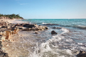 Rocky beach view at Caribbean sea coast, with light blue sky, turquoise water and white clouds. Sunny beach landscape with rocks and huge foamy waves at the ocean. Panoramic tropical landscape.