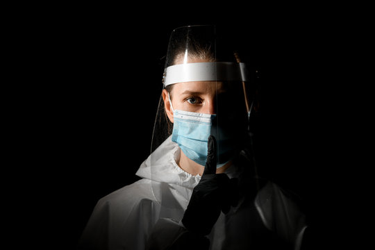young female nurse in medical mask and protective shield shows gesture of silence.