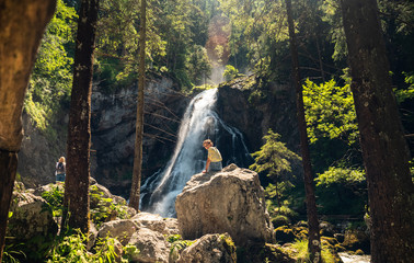 Young girl sitting on a rock in front of a waterfall in mountain forest with beautiful sun and lensflare