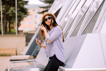 young girl with long brunette hair in straw hat with sunglasses listen music on her grey smartphone with headphones 