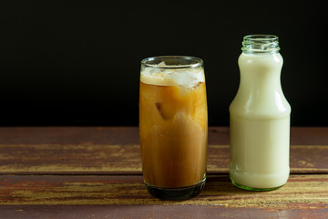 Ice coffee with milk and topping foam in glass and fresh milk in the glass bottle on old wood table and black background.