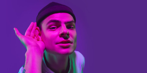 Attented listen with hand on ear. Caucasian man's portrait isolated on purple studio background in pink neon light. Beautiful male model. Concept of human emotions, facial expression, sales, ad.