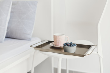 Obraz na płótnie Canvas Light breakfast served on a metal tray. Coffee in a pink cup and fresh blueberry on a white chair near bed. Coffee in bed concept