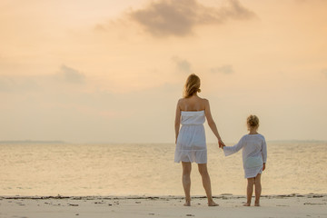 Back view of young mother and her adorable cute little girl holding hands and looking at beautiful ocean sunset. Happy family concept.