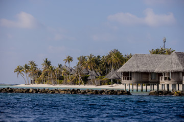 Landscape with exotic round tropical island with sandy beach, in background white clouds, houses and green palms, Indian ocean