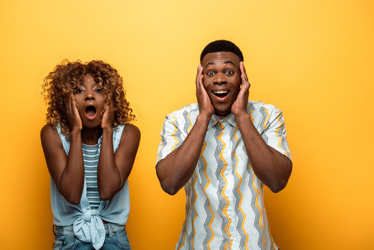shocked african american couple with open mouths touching faces on yellow colorful background
