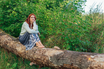 Romantic happy woman dressed in white sweater sitting on the dry fallen tree with bare feet