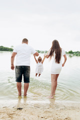 Mom, dad hugging daughter walking on the beach near lake. The concept of summer holiday. Mother's, father's, baby's day. Family spending time together on nature. Family look. Sun light.