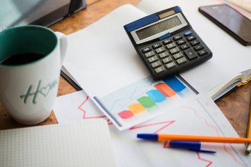 On the home table are stock and finance charts with a calculator, a coffee mug, a pencil, a marker pen, two notebooks, a smartphone and a tablet for remote work. Work at home.