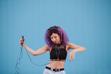 Ethnic teen woman listening to pop music with mobile phone. Technology and urban youth concept. Horizontal photo of millennial woman dancing on the street isolated on blue background.