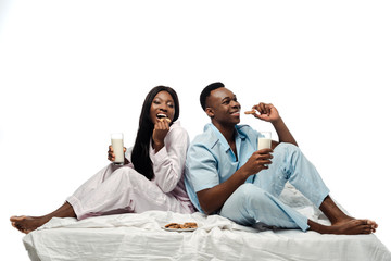 happy african american couple eating cookies with milk in bed in pajamas isolated on white