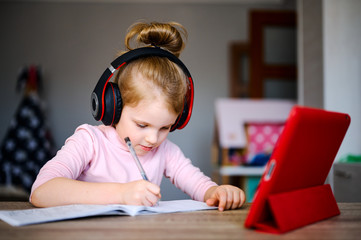 Distance learning online education. Schoolgirl in headphones studying at home with digital tablet and doing school homework. Training books and notebooks on table