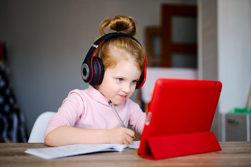 Distance learning online education. Schoolgirl in headphones studying at home with digital tablet and doing school homework. Training books and notebooks on table