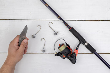 Fishing rod with a fishing reel and male hands with knife above white wooden background