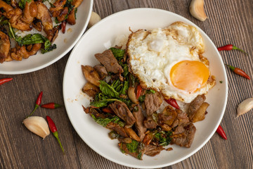 Pad Kra Pao Beef, (Stir fried beef with thai basil and chilli) top up with sun rise side up eggs on the white dish. close up , wooden table