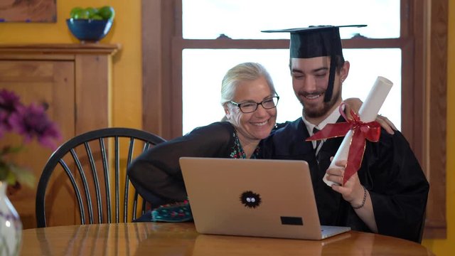 Young man graduate in costume along with mother at dining room table talk and show diploma to someone on laptop computer.